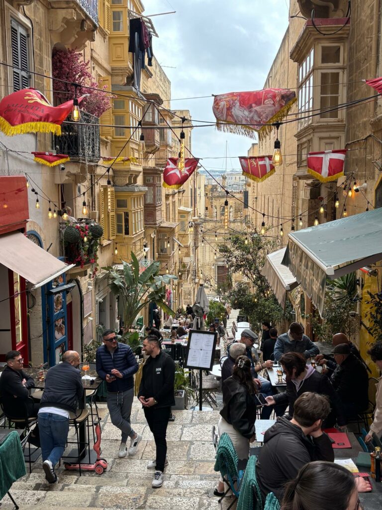 Pedestrian street full of people and bunting strung between buildings with the Maltese flag on it